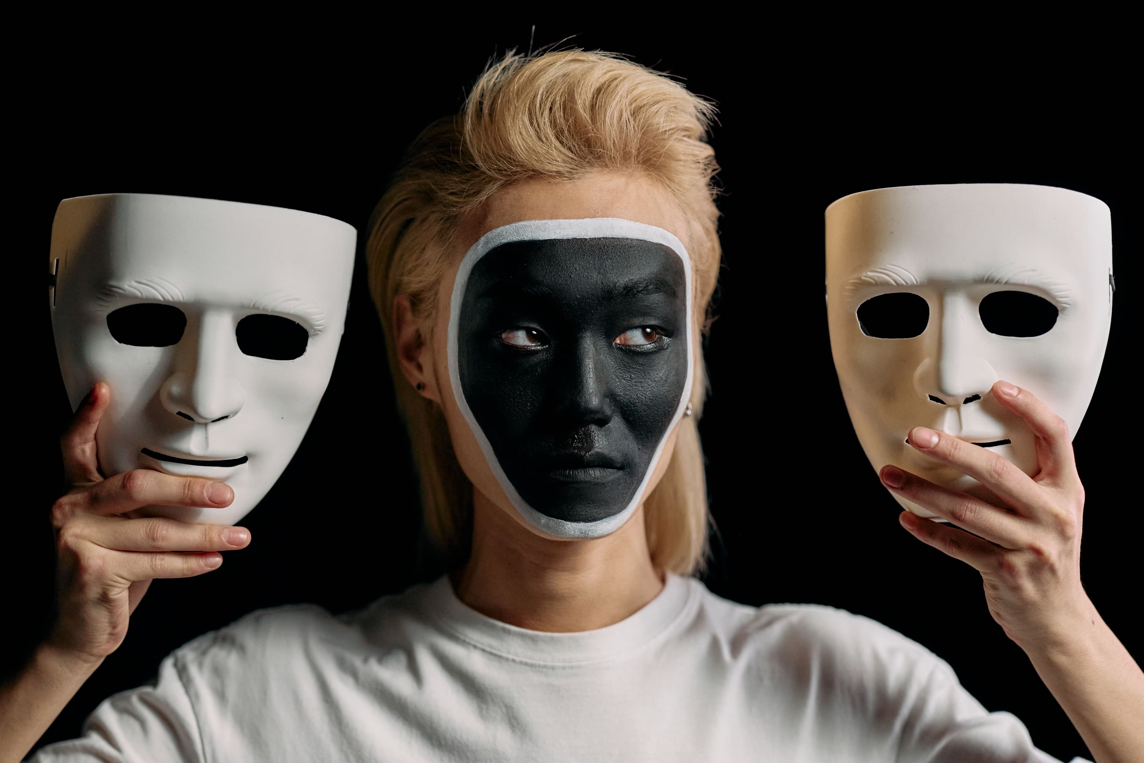 Person wearing a dark face covering while holding two white masks against a black background.